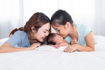 Obraz na płótnie Canvas Beautiful Asian mother and daughter spend time with newborn in bed, two woman kiss adorable infant with love and care. Toddler baby sleeping in bedroom with family.