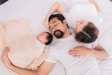 Asian young mother father and newborn sleep on bed together. Parent take a nap with baby because tired nurturing baby. Little infant  deeply sleeping wrapped in thin white cloth with happy and safe.