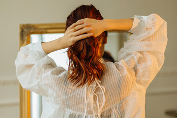 A beautiful red-haired girl in the bathroom looks in the mirror and ties her ponytail. Pretty woman...