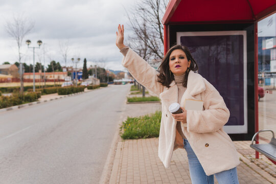 Young woman raising her hand to make the stop signal to the bus that comes on her route. Middle-aged girl at the bus stop with her hand up requesting public transportation.
