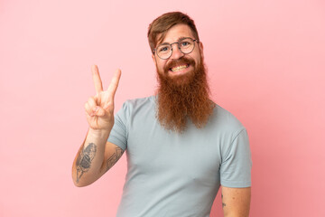 Young reddish caucasian man isolated on pink background smiling and showing victory sign