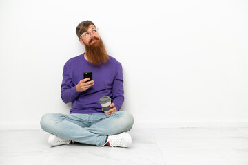 Fototapeta premium Young caucasian reddish man sitting on the floor isolated on white background holding coffee to take away and a mobile while thinking something