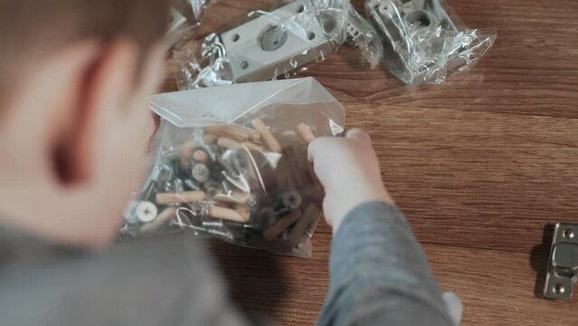 Boy examines set of fittings for assembling furniture in package. The child hands are touched and leveled by package with wooden dowels, eccentrics, bolts and nails, close-up. View from behind.