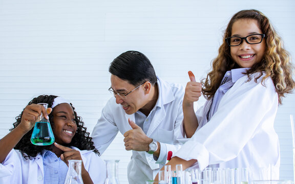 Asian handsome male teacher wearing eyeglasses and white gown uniform, teaching science and experiment to African black and Caucasian white female students in classroom at school. Education Concept.