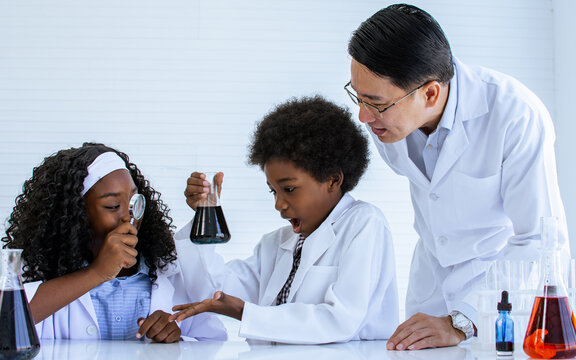 Asian handsome male teacher wearing eyeglasses and white gown uniform, teaching science and experiment to African black girl, boy students in classroom at school. Education Concept.