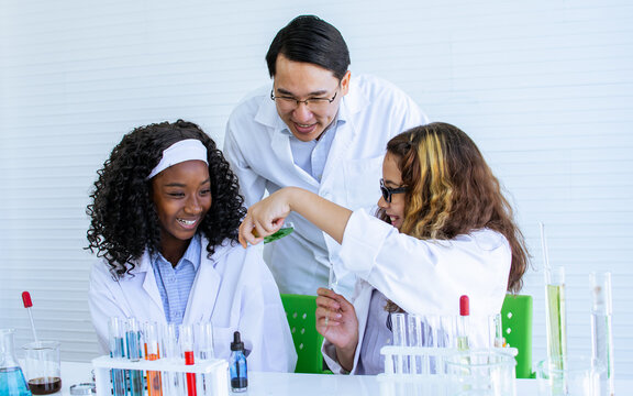 Asian handsome male teacher wearing eyeglasses and white gown uniform, teaching science and experiment to African black and Caucasian white female students in classroom at school. Education Concept.