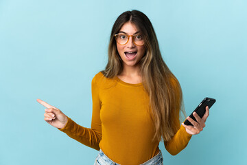 Young caucasian woman using mobile phone isolated on blue background surprised and pointing finger to the side