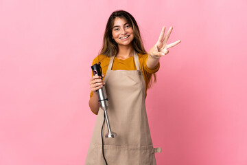 Young chef woman using hand blender isolated on pink background happy and counting three with fingers