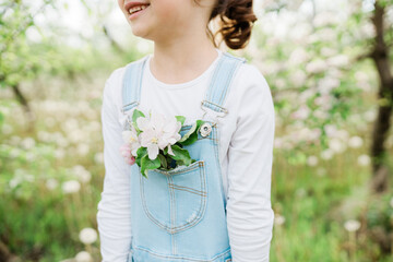 Obraz premium Little girl dressed in denim overalls, apple tree flowers in a pocket, blooming branch of an apple tree