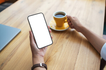 Mockup image of a woman holding mobile phone with blank desktop screen while drinking coffee