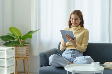 Smiling young Asian woman using tablet sitting on sofa at home.