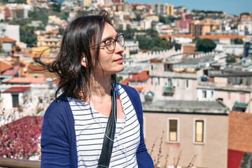 Fototapeta na wymiar Happy smiling woman listening music, song or audio guide at mobile phone while enjoying a view of panorama of Messina, Sicily, Italy. Urban traveling leisure.