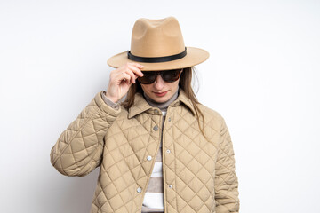 Fototapeta premium Young woman in glasses in a jacket with a hat lowering her head near a white wall.