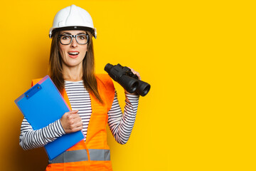 Young woman with a smile in a vest and hard hat holds a clipboard and binoculars on a yellow background