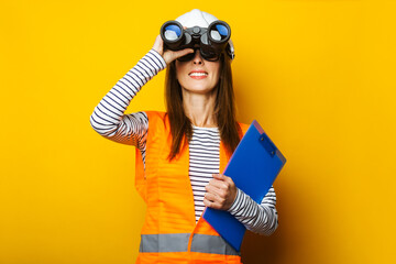 Young woman in signal vest and construction helmet looking through binoculars on yellow background
