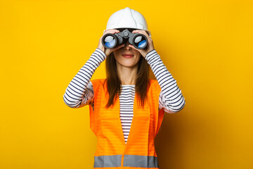 Young woman in a vest and a helmet looks through binoculars on a yellow background. Construction...