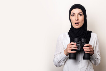 Friendly surprised Young Muslim woman in white shirt and hijab holds binoculars. Banner