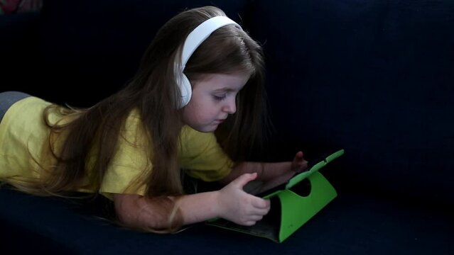 Cute small kid girl in headphones using tablet lying on sofa.