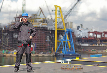 Inspector in coveralls and hardhat standing with mobile phone on harbor wharf. Shipyard in background.