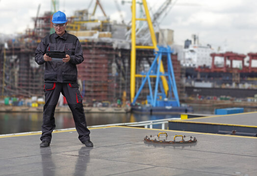 Inspector in coveralls and hardhat standing with tablet on harbor wharf. Shipyard in background.