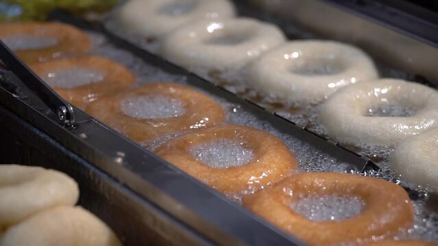 This video shows delicious hot and fresh donuts cooking in oil on factory conveyor belt on a busy day of food production.
