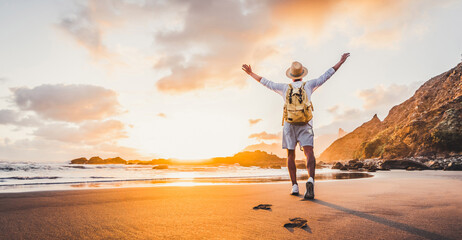 Happy man with hands up enjoying wellbeing and freedom at the beach - Male with backpack traveling in the nature with sunrise view - Healthy lifestyle, happiness and travel concept - obrazy, fototapety, plakaty