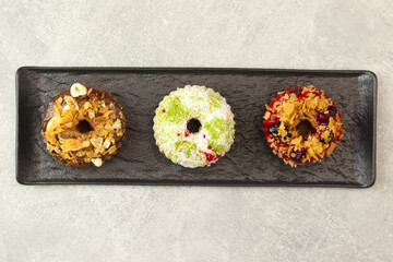 Vegan donuts topped with healthy red glaze with blueberry and flakes, coconut flakes and desiccated coconut, chocolate glaze with flakes, cocoa and nuts on black plate isolated on gray background.