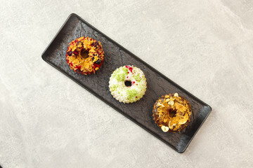 Vegan donuts topped with healthy red glaze with blueberry and flakes, coconut flakes and desiccated coconut, chocolate glaze with flakes, cocoa and nuts on black plate isolated on gray background.
