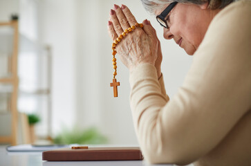 Elderly woman offering prayer. Humble religious senior lady sitting at table with Holy Bible,...