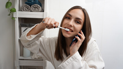 Busy young woman brushing teeth and talking on the phone simultaneously in front of a mirror. Morning bathroom rush concept.