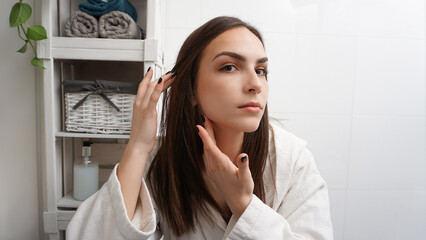Young woman in a bathrobe checking her face neck and lymph nodes in a bathroom mirror. Disease...