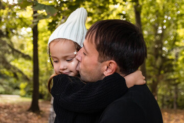 Fototapeta premium Dad and daughter hug and dad kisses daughter on the cheek. Summer park. Green trees. The concept of a young family. The decoration with stars is woven into the girl's hair.