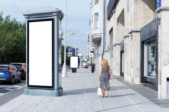 Tall Vertical Billboard On The Sidewalk. A Woman Is Walking With A White Bag. Mock-up.