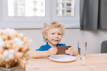 A little boy sits at a table and holds a bar of chocolate in his hands, looks at the camera, he is happy. White curly hair, wearing a blue t-shirt.