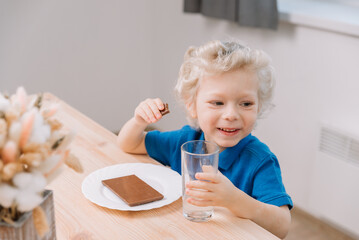 Obraz na płótnie Canvas A child boy sits at a table and eats a chocolate bar, he is happy and smiles, his gaze is directed to the side.