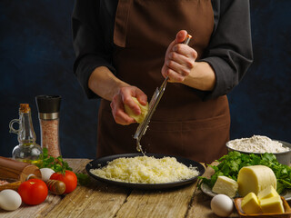 Preparation of dishes with grated cheese - pasta, pizza, risotto, salad. Chef rubs cheese on a manual grater on a dark blue background. Vegetables, herbs, eggs, spices on a wooden table.