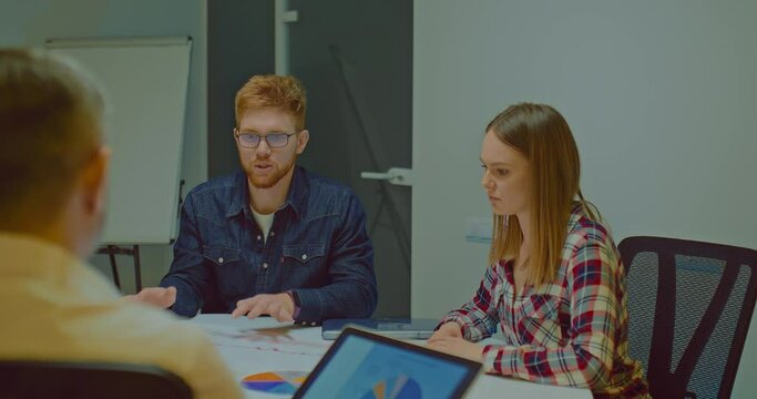 Red-haired businessman in glasses tells the company's development plans. Young entrepreneurs are discussing sales levels, charts of indicators are on the table. 4k, ProRes