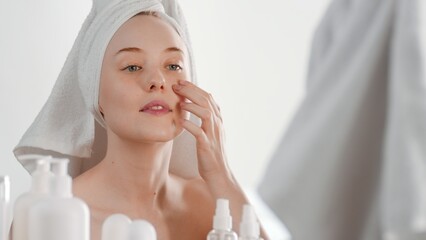 Beautiful young Caucasian woman with a towel on her head applies skin care cream on her cheek in...