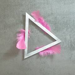 Pink feathers around a silver triangular frame on a gray concrete background. Text space. Top view. Minimal style.
