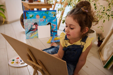 Beautiful European little girl enjoying learning painting, sitting on the floor at a wooden easel against the background of colorful joyful children painted images. Art class, creativity and education