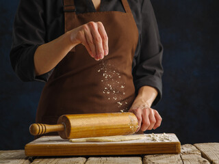 The chef rolls out the dough with a rolling pin on a wooden cutting board on a dark blue background. Close-up. Concept - recipes for pizza, pie, pasta, noodles. Restaurant, hotel, pizzeria.