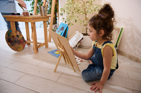 Concentrated European little girl enjoying painting, sitting on the floor at a wooden easel against the background of colorful joyful children painted images. Art class, creativity and education