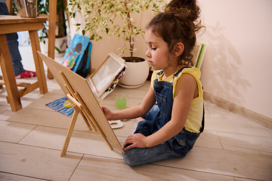 Beautiful girl, adorable preschool child, learns painting on canvas with watercolor or oil paints. Talent kid sits in front of wooden easel and enjoys art class at home. Education, creativity concept
