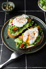 Toast with poached egg, lettuce, tomato and capers on a dark background, close up. Soft, selective focus