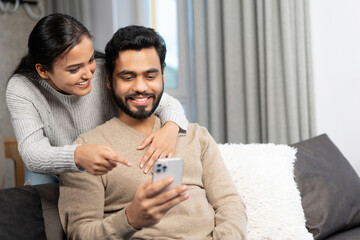Cheerful couple spends time with smartphone at home. Wife and a husband looks on the screen with smiles sitting on the couch at cozy living room, watching videos, scrolling feed and laugh