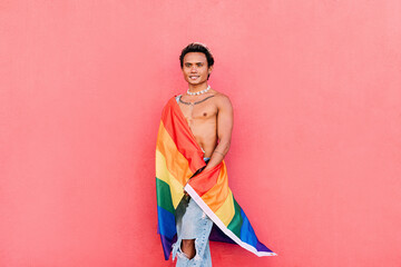 Young stylish man covered by rainbow LGBT flag leaning pink wall