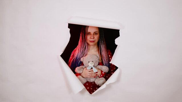 Young woman with teddy bear looking out of hole of white background. Pretty female with plush toy in hands.