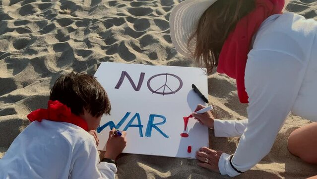 a mother and her son paint a sign on the sand on the beach that reads "no war". concept of pacifism and activism.