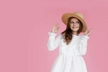 Front portrait of a cheerful little girl in elegant white dress, straw hat, looking at the side, on a pink background.