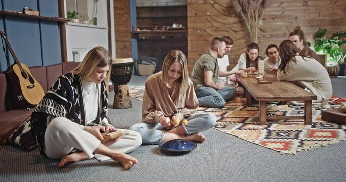 Female hippie playing music together. Young girlfriends playing tank drum and kalimba during meeting with friends in spacious loft room
